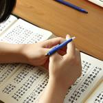 **Prompt:** A focused individual, likely a young adult, immersed in writing Korean characters in a traditional notebook with a pen. Surrounding them are scattered Korean language textbooks, flashcards, and a smartphone displaying a language learning app. The scene should evoke a sense of deep personal engagement and dedicated effort. Soft, warm lighting highlights the meticulous act of writing, symbolizing the human touch, emotional investment, and the journey of mastering Korean script and culture. The background hints at a cozy, studious atmosphere, possibly with subtle Korean cultural elements.